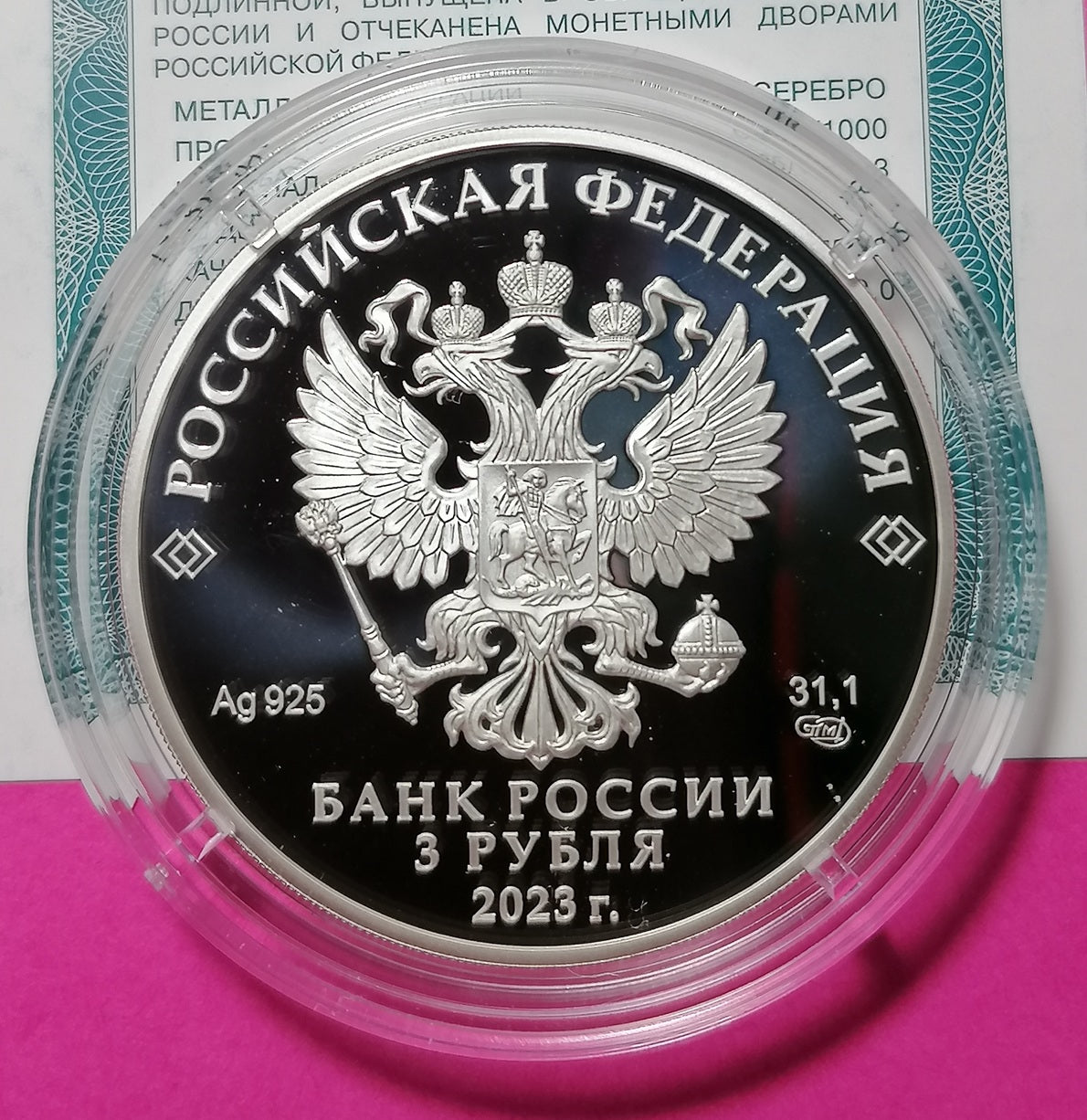 A silver Russian 3 ruble coin from 2023, featuring the double-headed eagle coat of arms, encased in a clear protective plastic capsule. The coin is displayed against a background containing a Russian certificate and a bright pink surface.